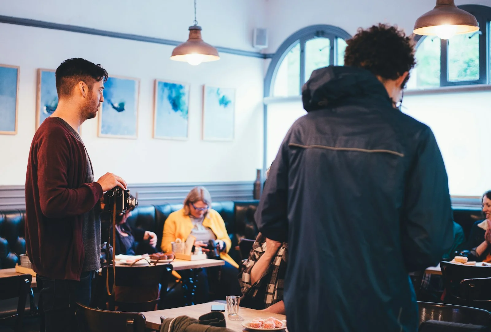 Man holding a vintage camera stands in a busy small café as customers chat at tables