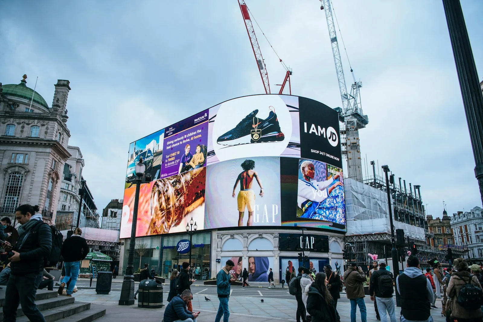 Pedestrians gather beneath giant curved digital billboards showing GAP and JD adverts beside construction cranes