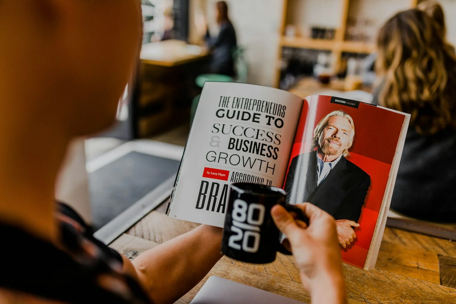 Person holding an 80/20 mug reads a magazine feature on business growth in a café